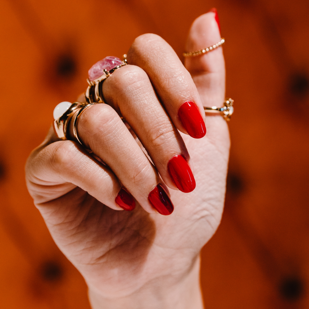 Classic Red Elegant Manicure on white female with gold rings and an orange background.