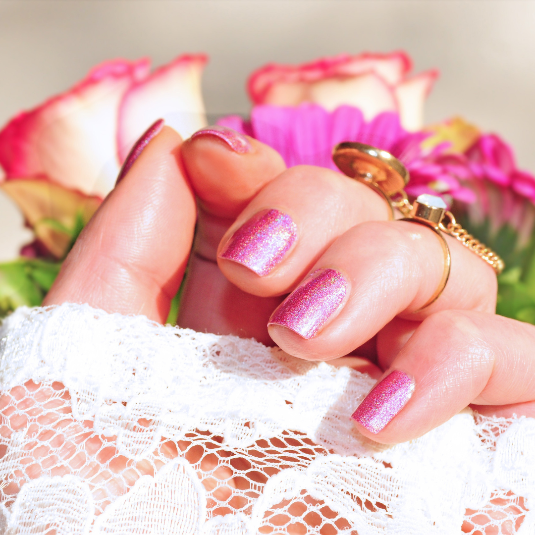 Pink metallic manicure on a female hand with white lace sleeve showing and blurry roses as the background.