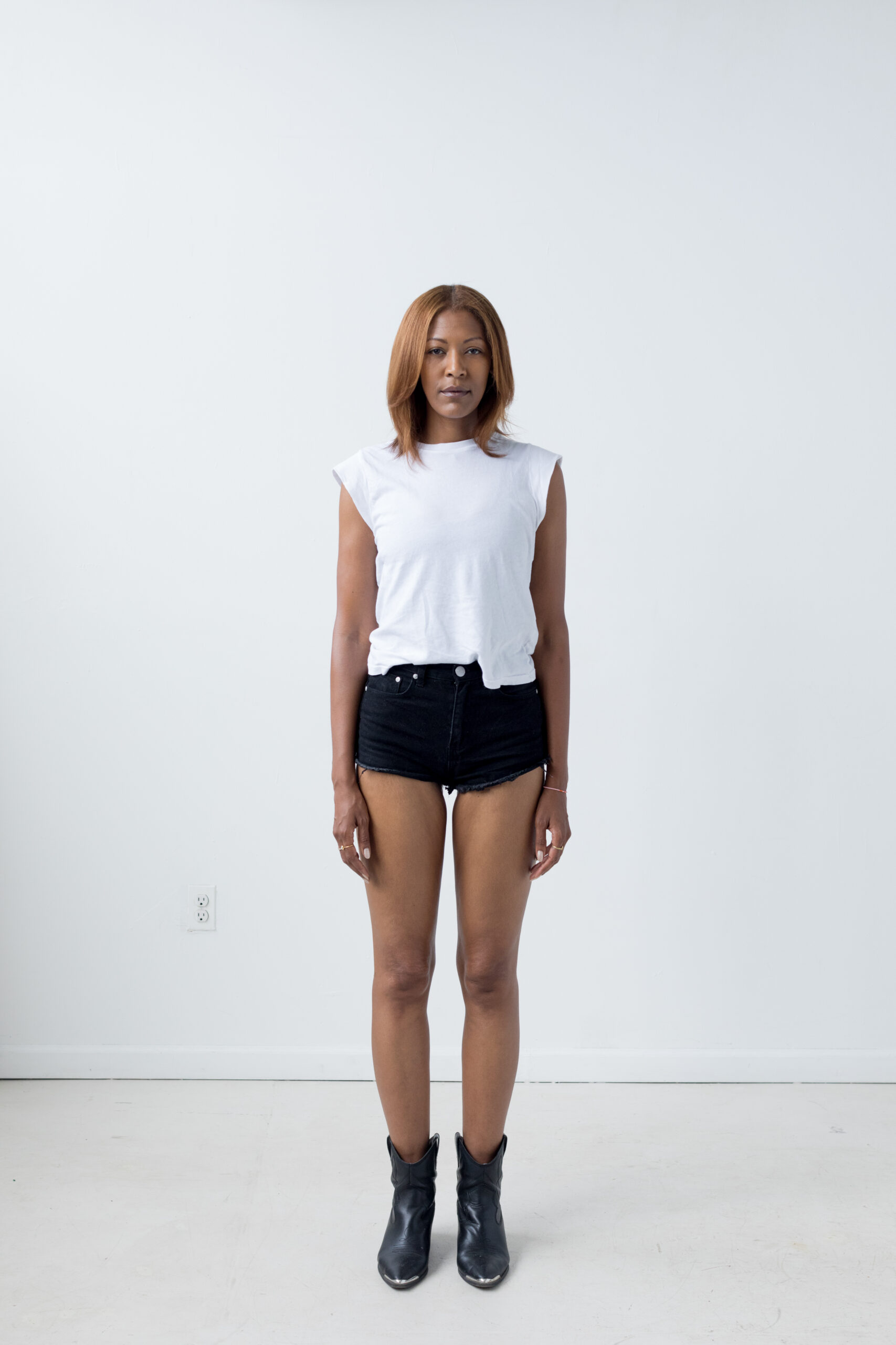 The model stands tall with feet together and arms by her side, against a white backdrop. Model is wearing black denim shorts with a white sleeveless top.